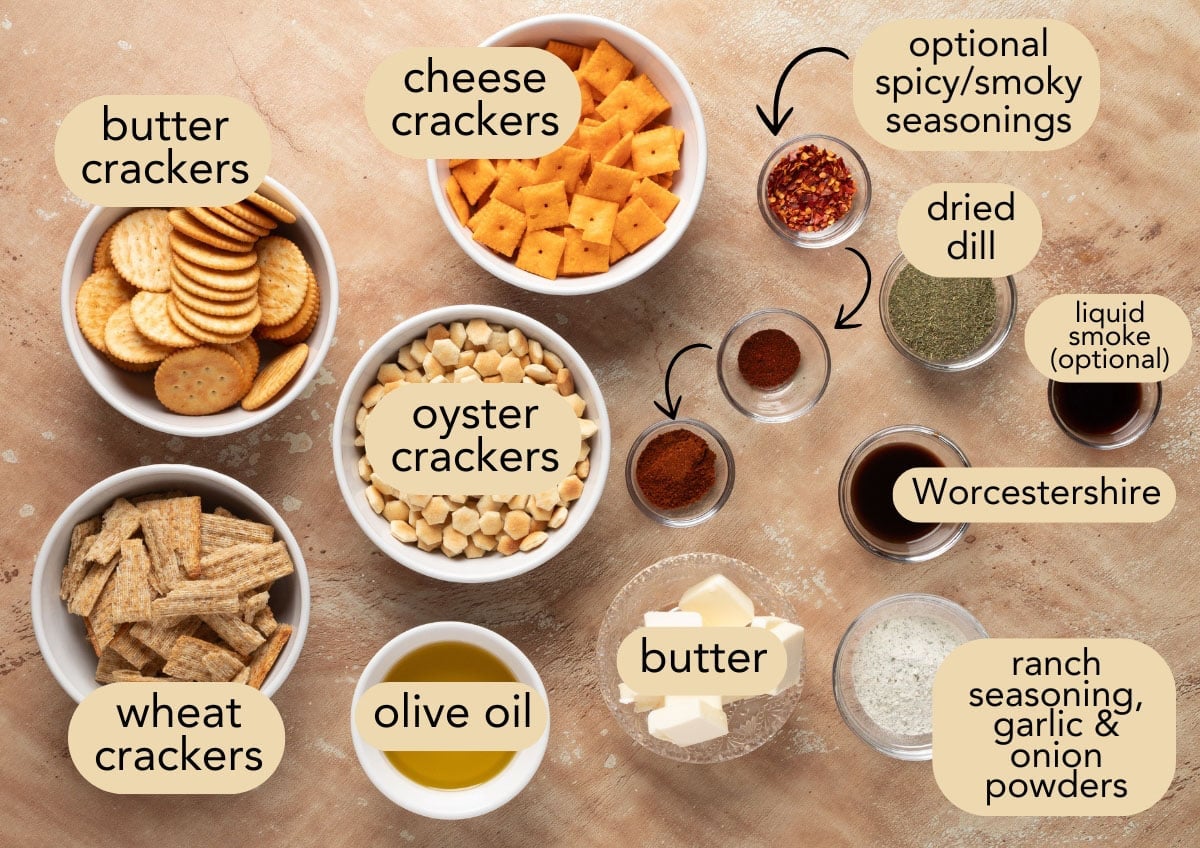 Flat lay of labeled ingredients for Dill Ranch Snack Mix on a beige surface. Bowls of butter crackers, wheat crackers, cheese crackers, and oyster crackers are arranged with olive oil, butter, Worcestershire sauce, ranch seasoning, dried dill, garlic and onion powders, and optional smoky or spicy seasonings, including red pepper flakes, chili powder, and liquid smoke.