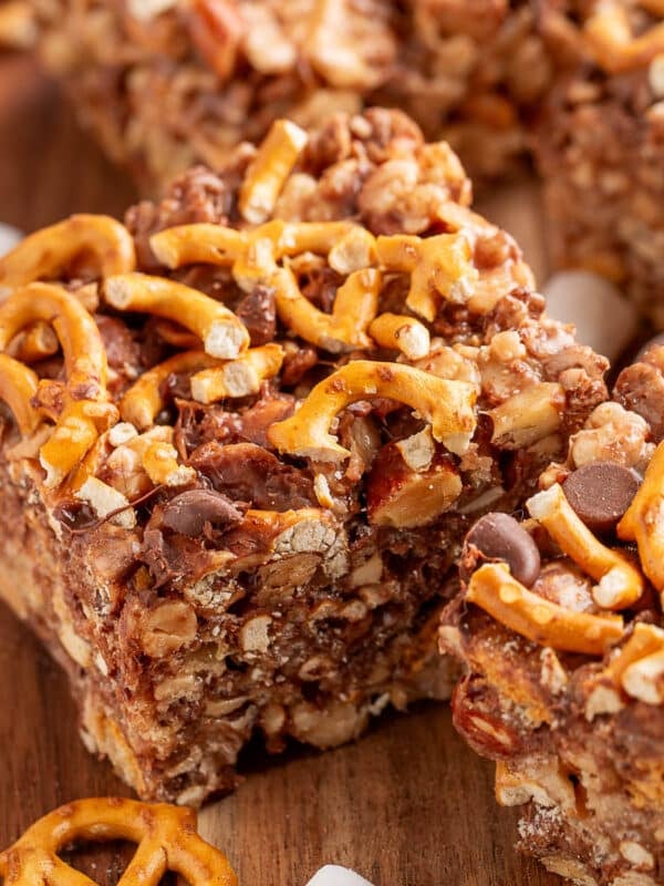 Close-up of kitchen sink rice krispie treats cut into squares on a wooden board. The bars are packed with pretzel twists, chocolate chips, nuts, and marshmallows, creating a sweet and salty texture. Pieces of broken pretzels and mini marshmallows are scattered around the board.