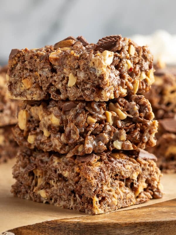 Stack of three peanut butter cup rice krispie treats on a wooden board lined with parchment paper. The bars are thick and chewy, filled with chocolate puffed rice cereal, chopped peanuts, and bits of peanut butter cups. A few mini marshmallows and peanut butter chips are scattered at the bottom of the frame.