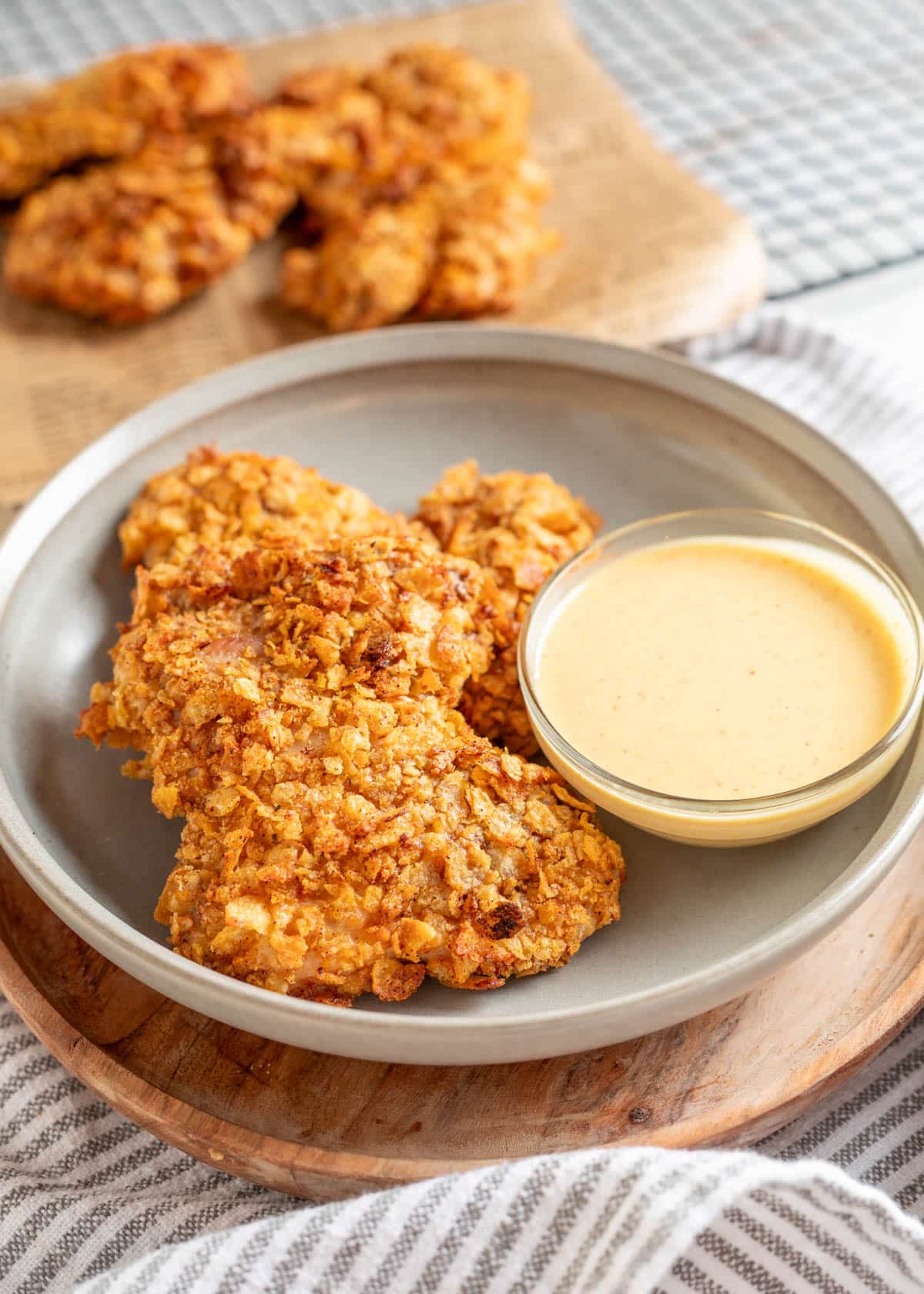 Crispy potato chip&ndash;crusted chicken thighs served on a gray plate with a small bowl of creamy dipping sauce. More pieces of chicken are visible on parchment paper in the background.