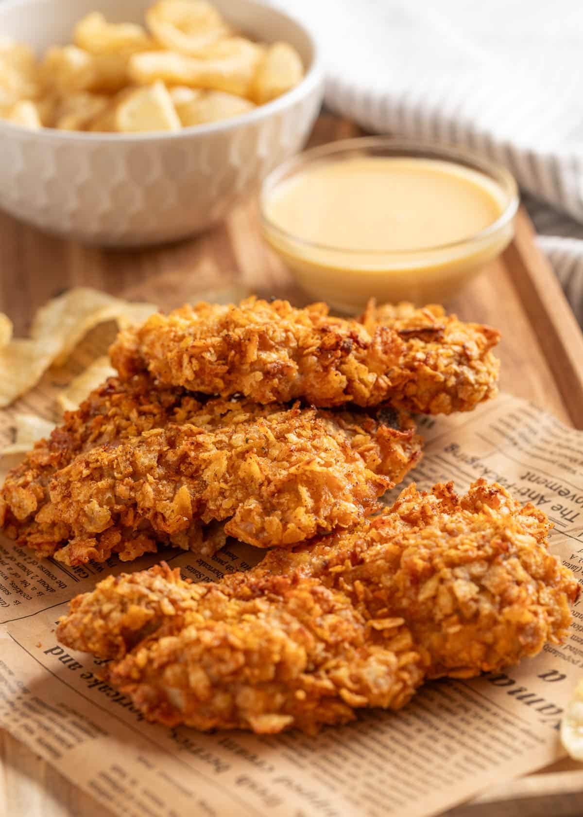 Crispy potato chip&ndash;crusted chicken thighs on a tray lined with faux newsprint, served with a small bowl of honey mustard dipping sauce and a side of kettle-style potato chips in the background.