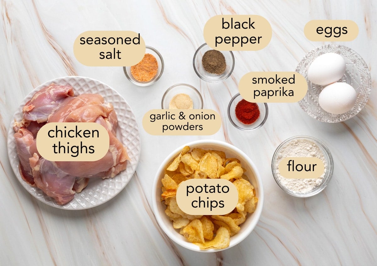 Ingredients for Air Fryer Potato Chip Chicken arranged on a white marble surface, each labeled with beige text bubbles. Shown are raw boneless chicken thighs on a plate, a bowl of crushed potato chips, a bowl of flour, two eggs in a glass dish, and small bowls of spices including seasoned salt, garlic & onion powders, black pepper, and smoked paprika.
