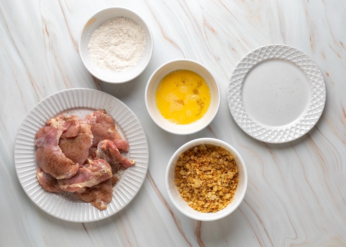 A top-down view of a marble countertop setup for breading chicken thighs. A plate of raw, seasoned chicken thighs sits at the lower left. Around it are three bowls containing flour, beaten eggs, and crushed potato chips, plus an empty plate at the top right for the breaded chicken.