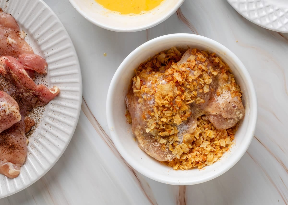 Chicken thighs coated in egg and being pressed into a bowl of crushed potato chips. The chip coating clings to the chicken with visible seasoning. Part of the raw chicken plate and egg wash bowl are visible nearby.