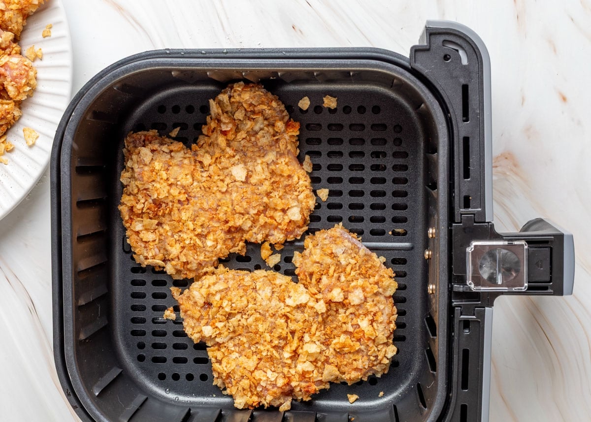 Two pieces of potato chip-coated chicken arranged in a black air fryer basket, ready to cook. More breaded pieces are visible on a white plate just outside the basket.