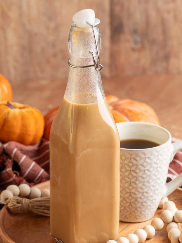 Glass swing-top bottle filled with creamy pumpkin spice coffee syrup on a wooden tray, next to a patterned white mug of black coffee. Decorative pumpkins, a striped napkin, and a wood bead garland add a fall-inspired touch.