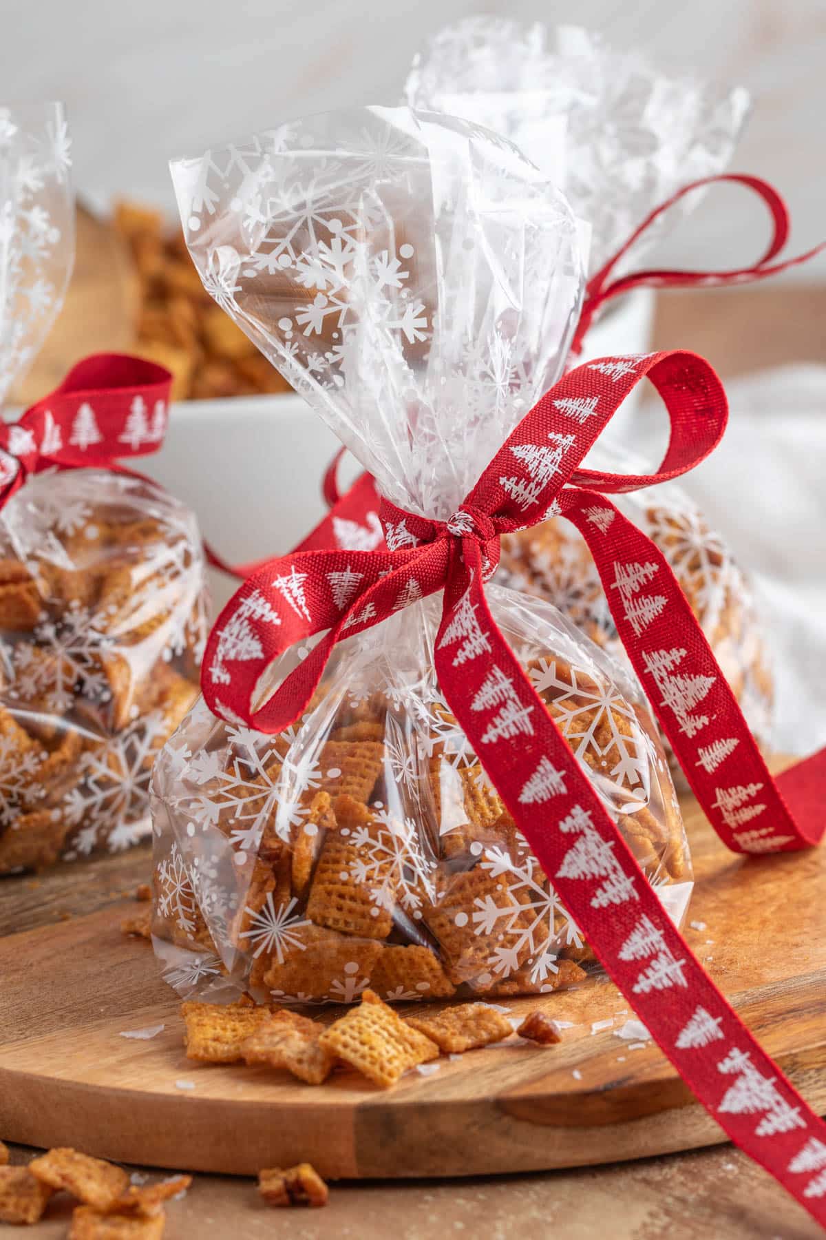 Clear cellophane gift bags filled with homemade snack mix, tied with red ribbon printed with white Christmas trees. The bags sit on a wooden board with scattered snack pieces around them.