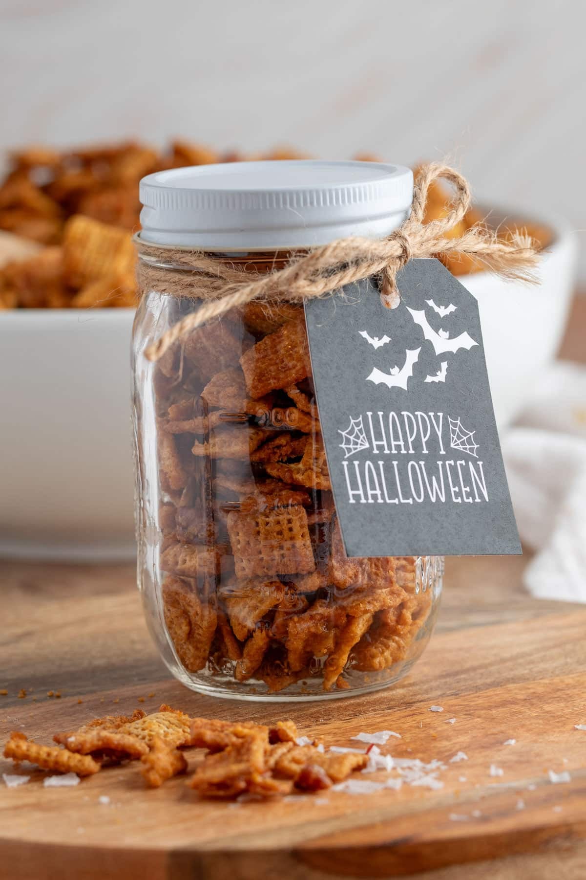 Glass jar filled with cinnamon praline snack mix, tied with twine and adorned with a black &ldquo;Happy Halloween&rdquo; gift tag featuring bats and spiderwebs. A bowl of snack mix is blurred in the background.