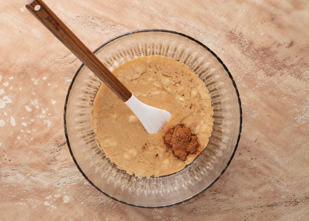 Overhead view of a glass mixing bowl filled with a bubbly butter and sugar mixture, with a mound of cinnamon on one side. A white spatula with a wooden handle rests in the bowl, ready to stir.