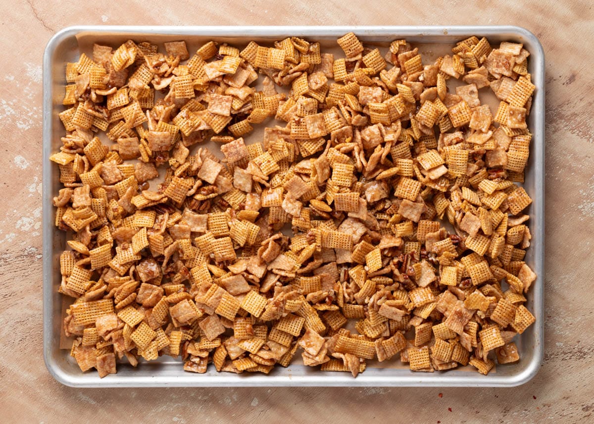 Cinnamon Praline Crunch Snack Mix spread evenly on a parchment-lined baking sheet before baking; cereal pieces glistening with caramelized coating.