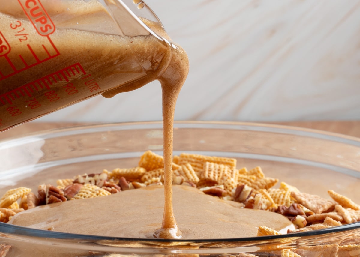 Close-up of cinnamon praline glaze being poured from a measuring cup over Chex cereal and pecans for a sweet and crunchy snack mix.