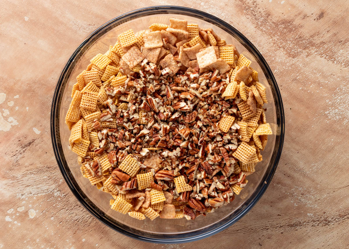 Top-down view of a clear glass mixing bowl filled with corn cereal squares, cinnamon toast cereal, and chopped pecans, on a tan textured surface.