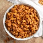 Overhead view of a large white bowl filled with cinnamon praline crunch snack mix, featuring golden Chex cereal coated in a caramelized praline mixture with chopped pecans, set on a rustic surface with a white linen napkin.