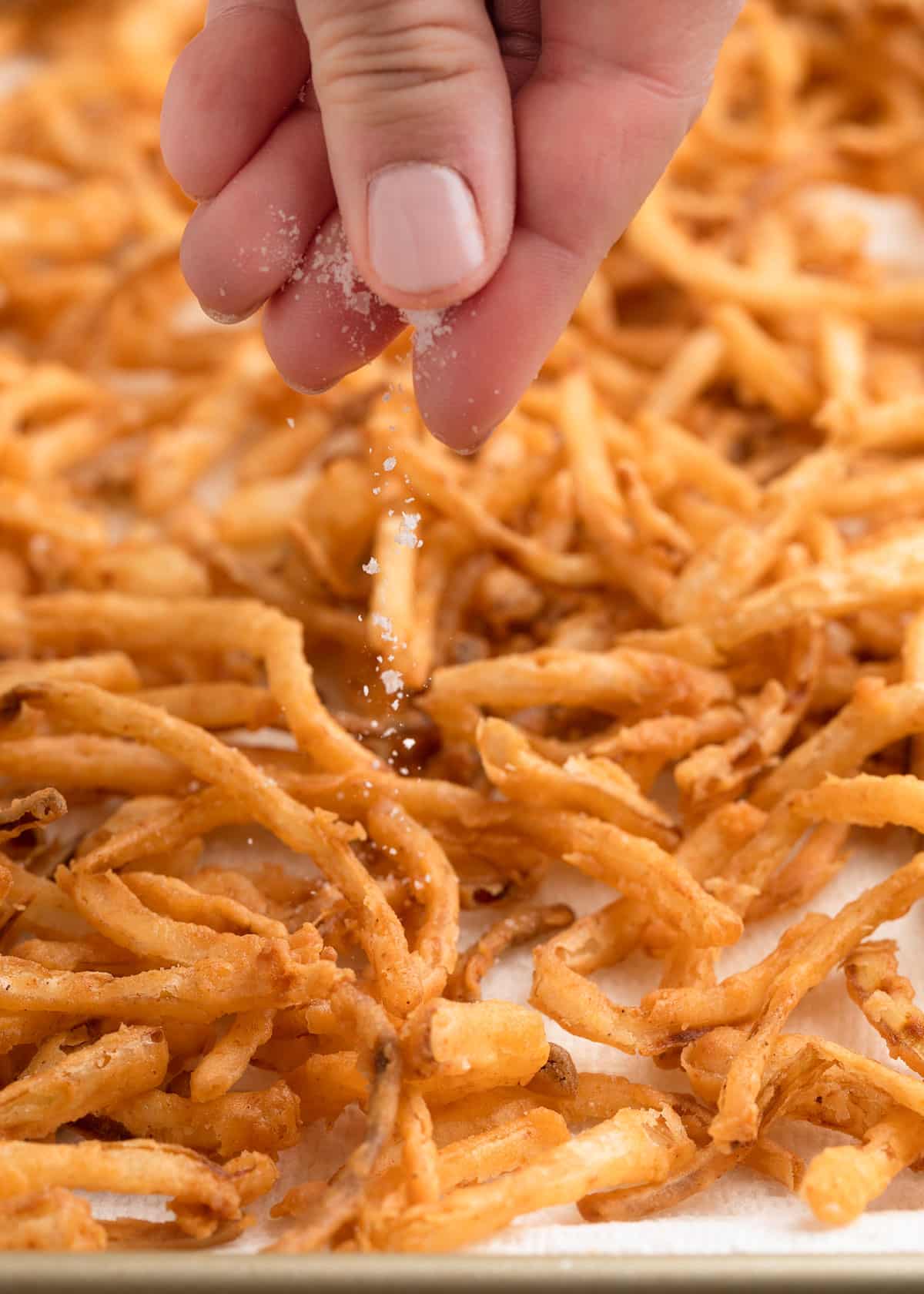 A close-up of a hand sprinkling salt over a tray of crispy homemade fried onions on parchment paper.