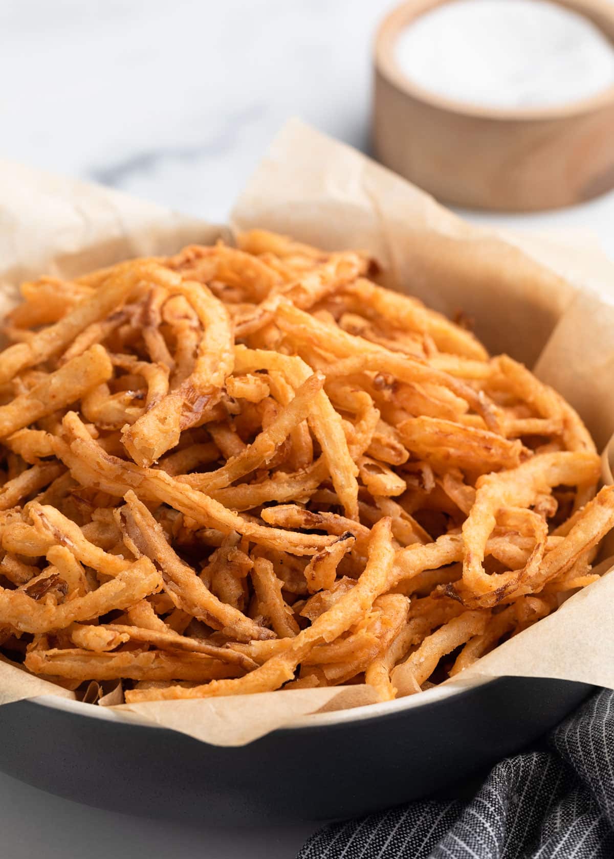 A close-up of a black bowl lined with parchment paper, filled with crispy homemade French fried onions, with a gray striped napkin and a wooden salt cellar blurred in the background.