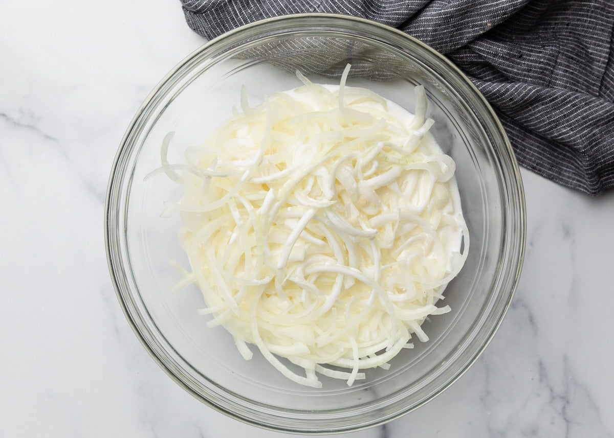 Sliced onions soaking in a glass bowl filled with buttermilk, placed on a marble surface with a gray striped towel nearby.