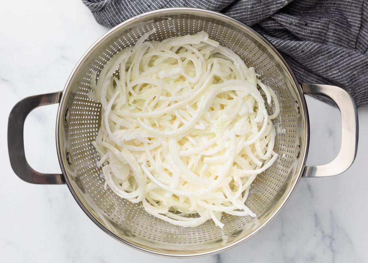 Sliced onions in a metal colander set over a marble surface, with a gray striped kitchen towel to the side.