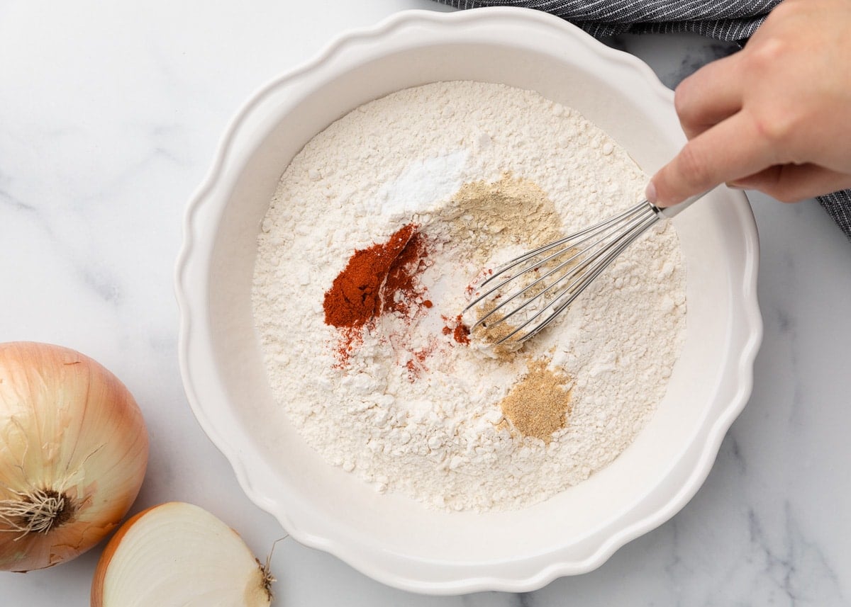 A white ceramic bowl filled with flour, baking powder, and spices, including paprika and garlic powder, with a hand whisking the seasoning blend together.
