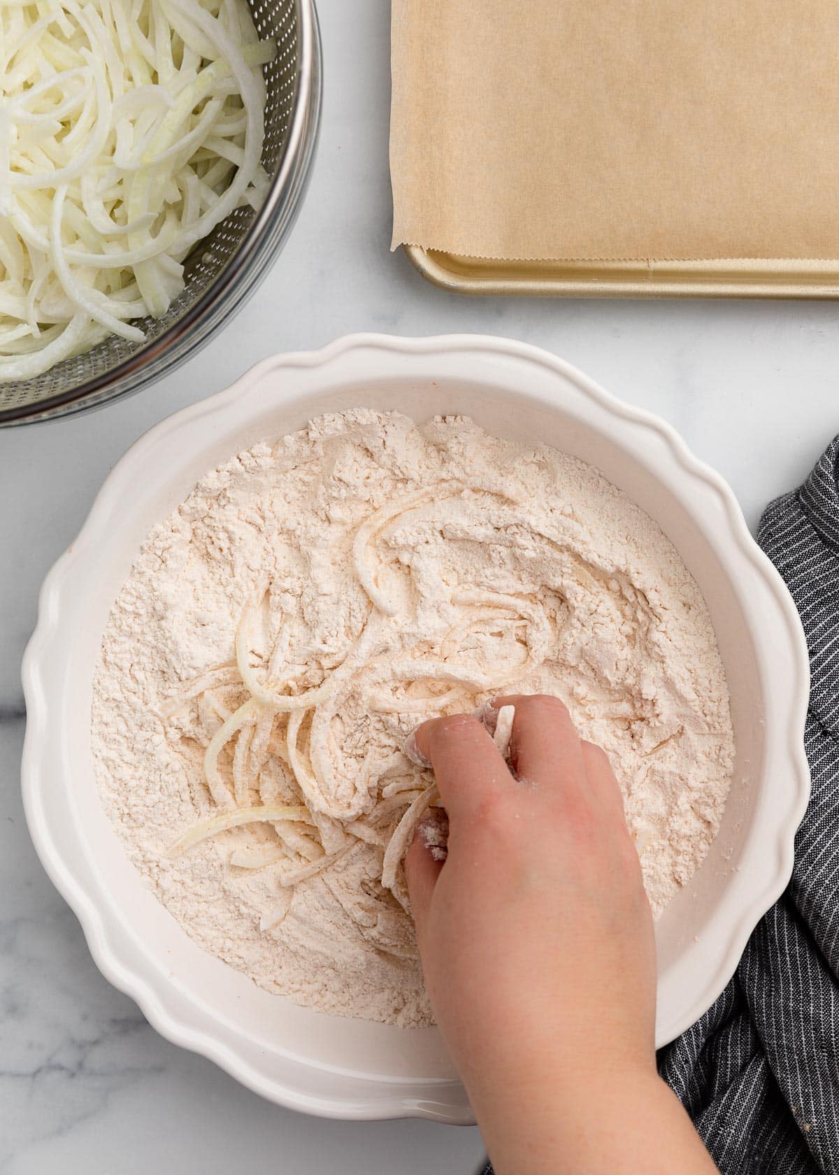 A hand tossing sliced onions in seasoned flour inside a white bowl, with a metal colander of sliced onions and a parchment-lined baking sheet in the background.