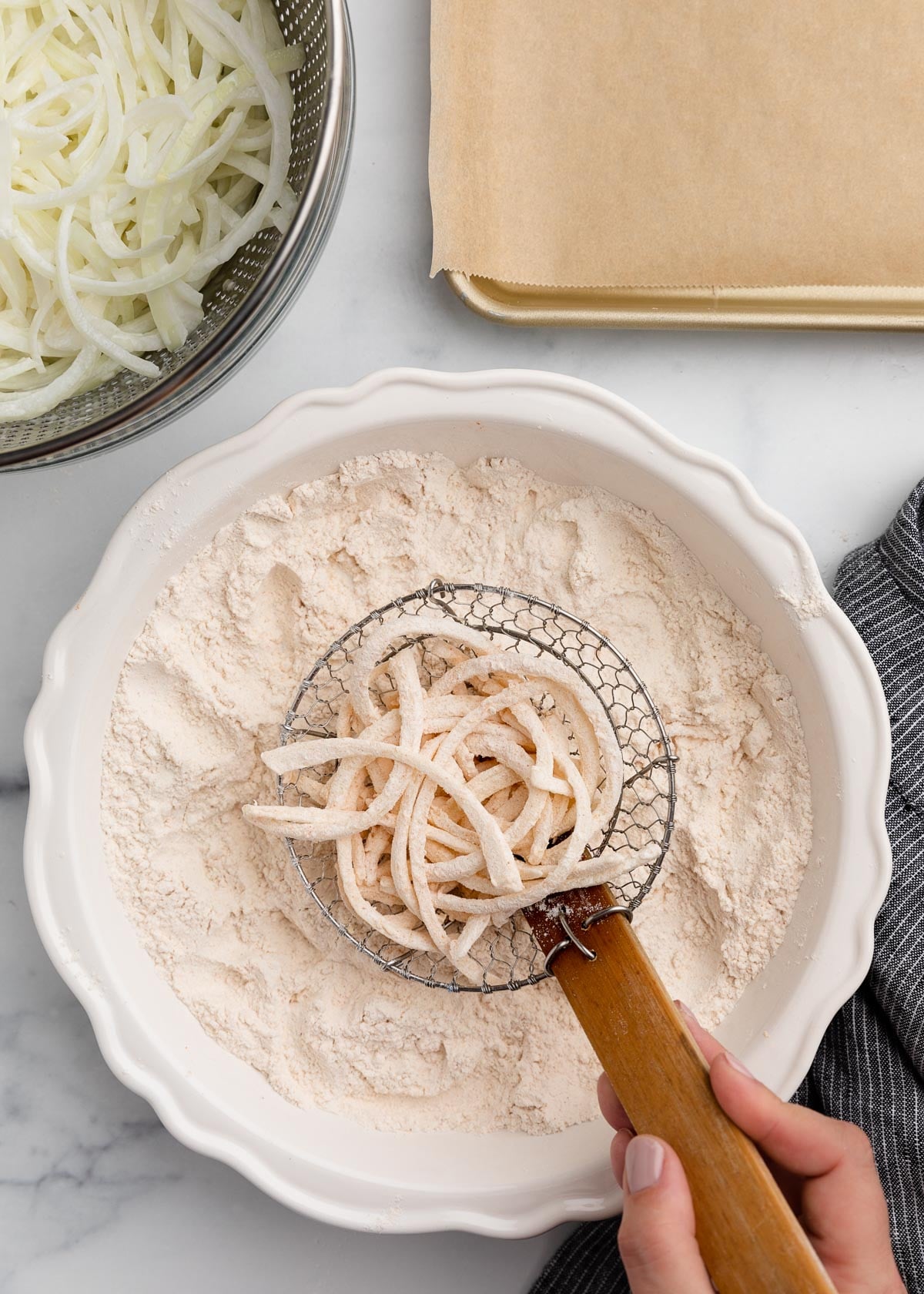 A wire spider strainer holding a portion of flour-dredged onion slices over a bowl of seasoned flour, next to a colander of sliced onions and a prepared baking sheet.