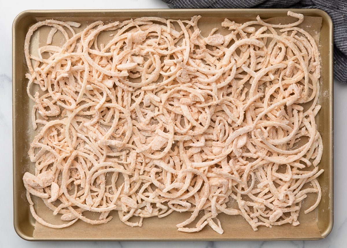 Thinly sliced onions coated in seasoned flour mixture, spread evenly on a parchment-lined baking sheet, ready for frying.