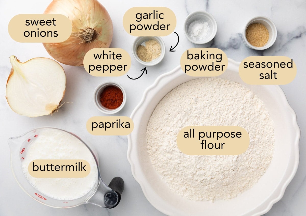 Labeled ingredients for homemade French fried onions arranged on a marble surface, including sweet onions (whole and halved), buttermilk, all-purpose flour, paprika, white pepper, garlic powder, baking powder, and seasoned salt.