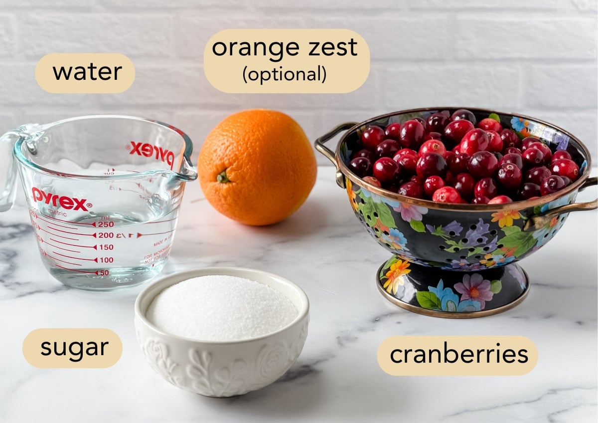 Overhead view of ingredients for homemade cranberry sauce, labeled with text: a glass measuring cup of water, a whole orange labeled &ldquo;orange zest (optional),&rdquo; a black floral colander filled with fresh cranberries, and a white bowl of sugar.