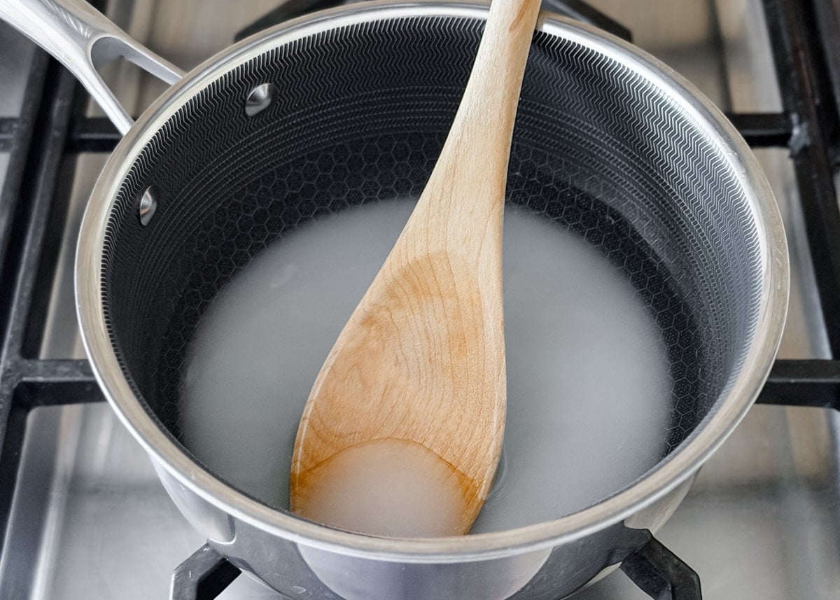 A saucepan on a stovetop with a wooden spoon stirring a cloudy mixture of sugar and water as it begins to dissolve.