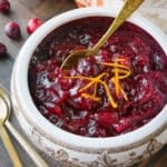 Overhead view of a white and brown patterned serving bowl filled with whole berry cranberry sauce, garnished with strips of fresh orange zest and a gold spoon; scattered cranberries and gold flatware rest on a dark wood surface in the background.