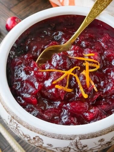 Overhead view of a white and brown patterned serving bowl filled with whole berry cranberry sauce, garnished with strips of fresh orange zest and a gold spoon; scattered cranberries and gold flatware rest on a dark wood surface in the background.