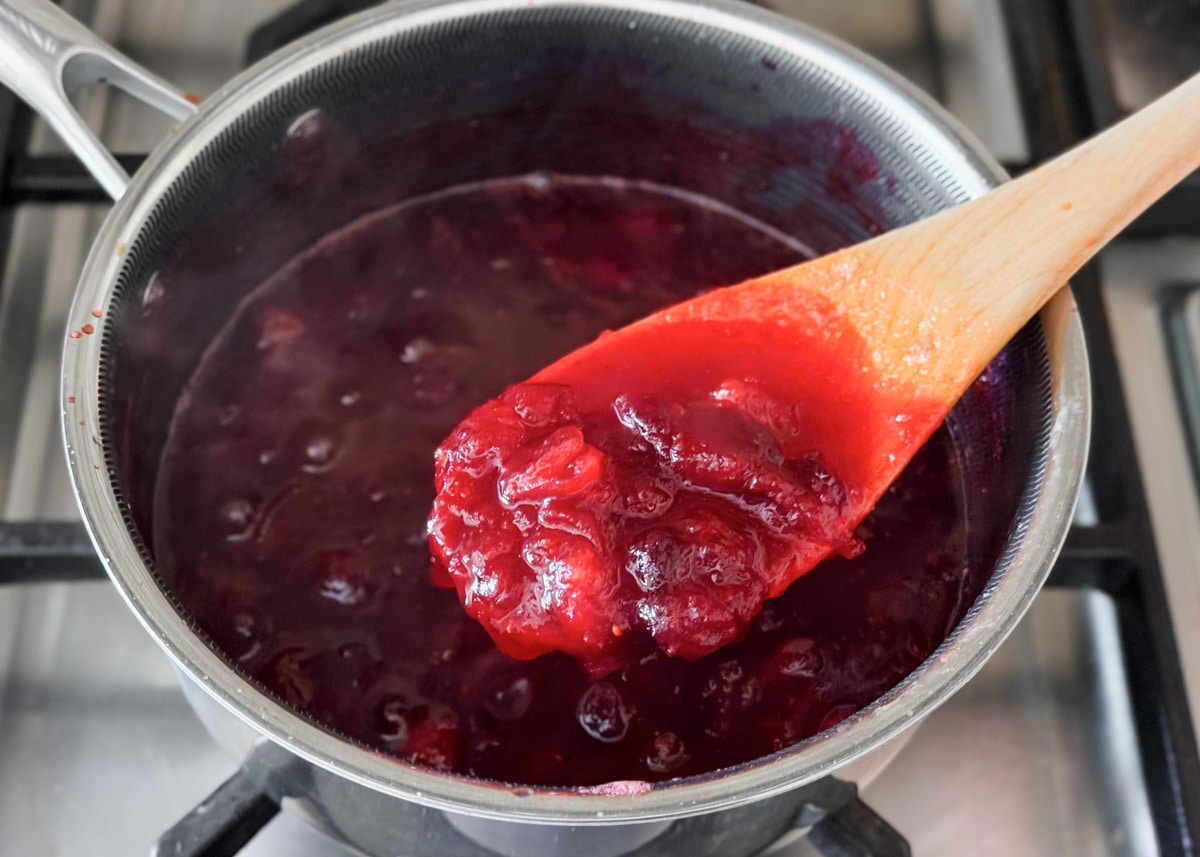 A close-up of thickened cranberry sauce in the saucepan, with a wooden spoon lifting a scoop to show texture.