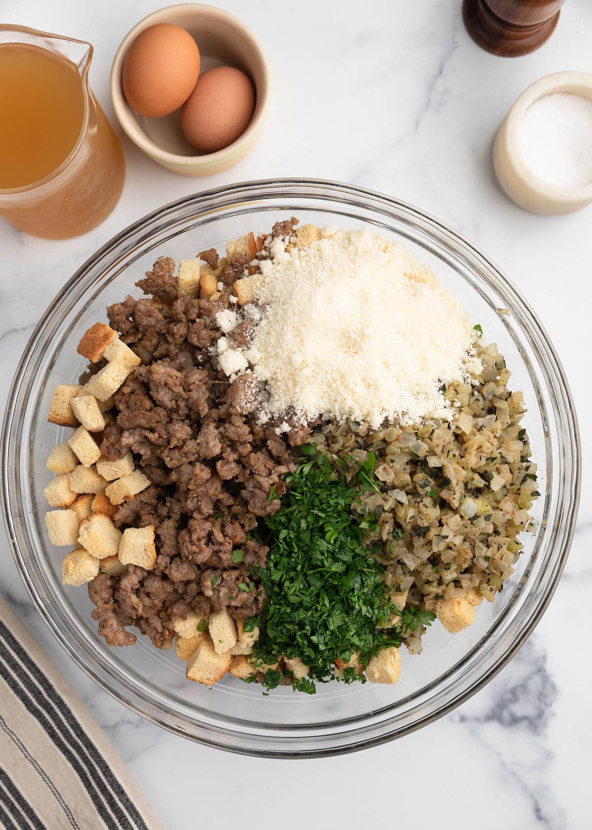 Overhead view of a glass mixing bowl filled with stuffing ingredients, including cooked Italian sausage, cubed bread, saut&eacute;ed onions and celery, chopped parsley, and grated cheese, surrounded by eggs, broth, and seasonings on a marble surface.
