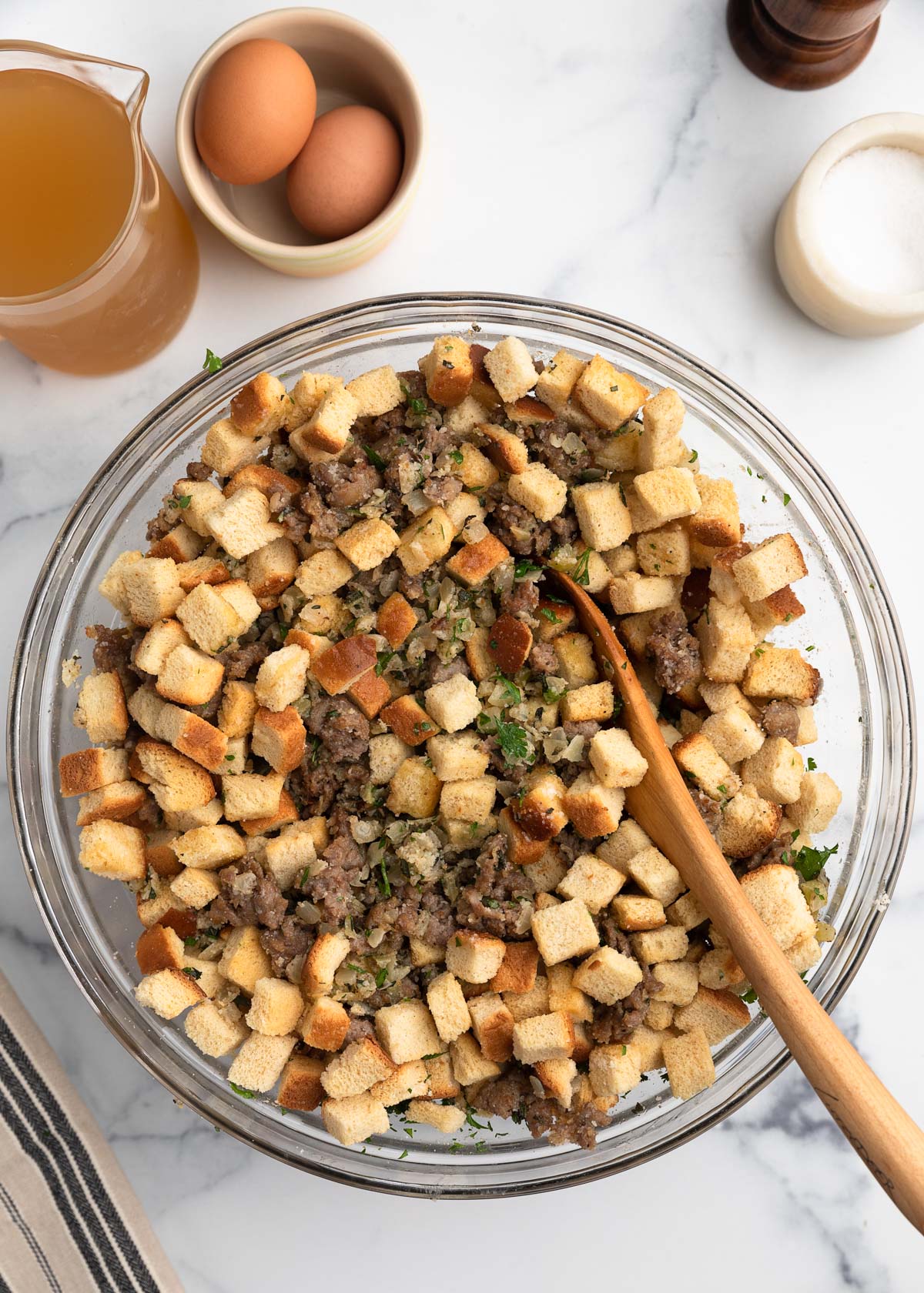 Overhead view of partially mixed Italian sausage stuffing in a glass bowl, showing cubed bread, sausage, herbs, and vegetables being combined with a wooden spoon, with eggs and broth nearby on the countertop.