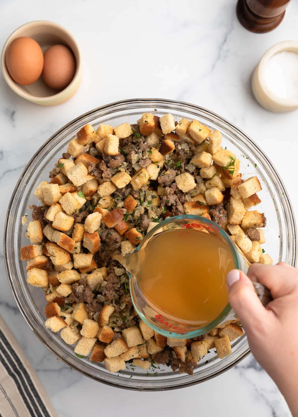 A hand pouring chicken broth from a glass measuring cup into a large glass bowl filled with cubed bread, cooked sausage, onions, and fresh herbs for stuffing.