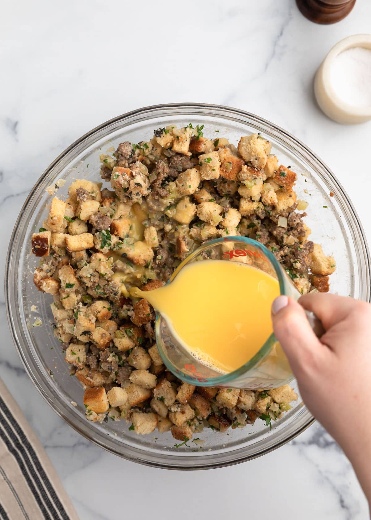 Beaten eggs and stock being poured from a measuring cup into a bowl of sausage stuffing mixture with bread cubes, herbs, and broth on a white marble surface.