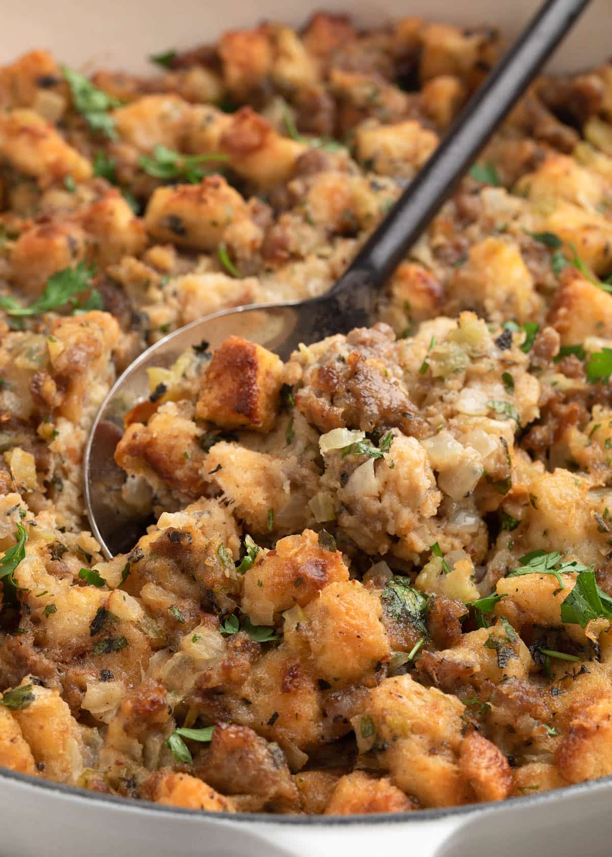 Close-up of a serving spoon scooping Italian sausage stuffing from a white dish, featuring browned bread cubes, crumbled sausage, onions, celery, and chopped parsley.