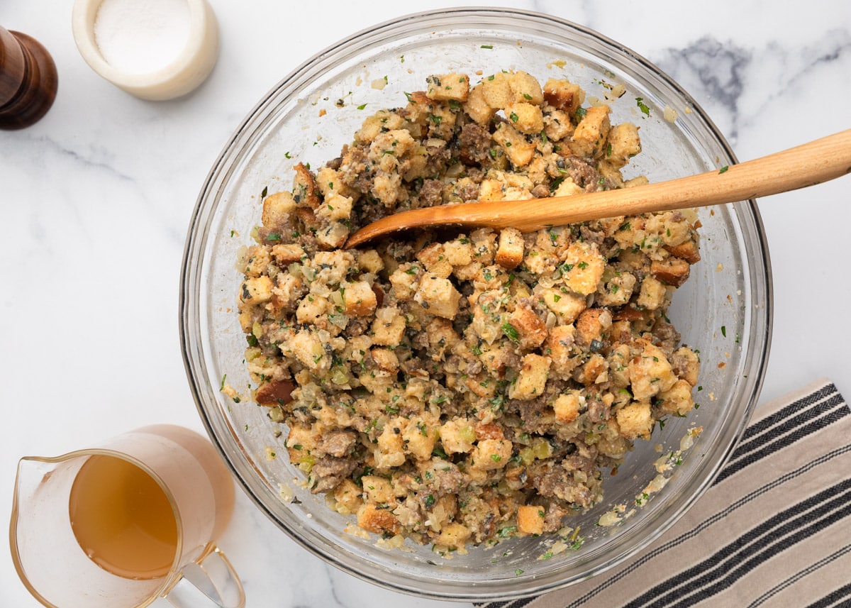 Overhead view of sausage stuffing mixture in a large glass mixing bowl with a wooden spoon. Chopped bread, sausage, celery, and herbs are visible. A pitcher of broth sits beside the bowl.