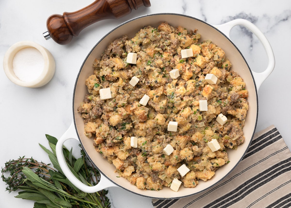 Overhead view of unbaked sausage stuffing in a white braiser, dotted with cubes of butter on top. Fresh herbs, salt, and pepper are arranged nearby on a marble surface.