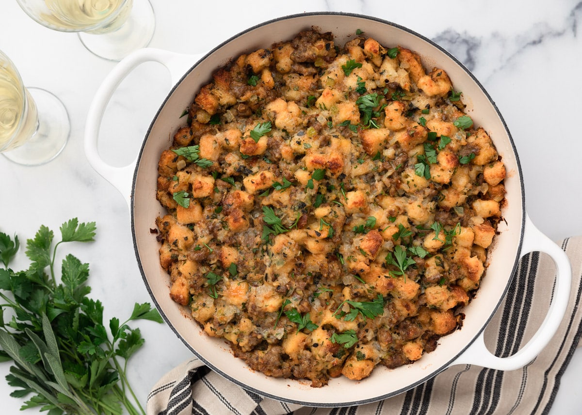 Overhead view of baked sausage stuffing in a white braiser, golden and crisp on top, garnished with fresh parsley. Glasses of white wine and fresh herbs are arranged nearby for serving.