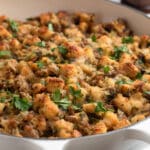 Golden-brown Italian sausage stuffing in a white braiser, topped with chopped fresh parsley. The stuffing features toasted bread cubes, crumbled sausage, and visible bits of onion and celery.