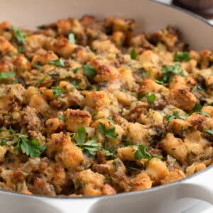 Golden-brown Italian sausage stuffing in a white braiser, topped with chopped fresh parsley. The stuffing features toasted bread cubes, crumbled sausage, and visible bits of onion and celery.