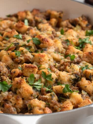 Golden-brown Italian sausage stuffing in a white braiser, topped with chopped fresh parsley. The stuffing features toasted bread cubes, crumbled sausage, and visible bits of onion and celery.