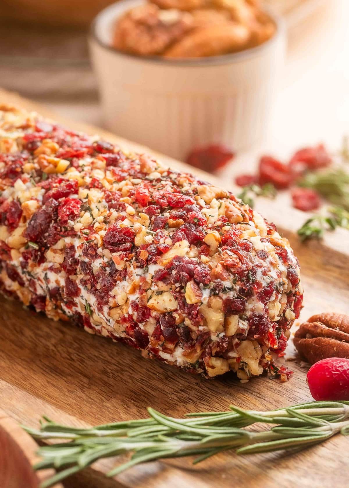 Close-up of a log-shaped goat cheese appetizer coated in finely chopped cranberries, toasted pecans, and fresh herbs, displayed on a wooden board with rosemary sprigs and whole pecans.