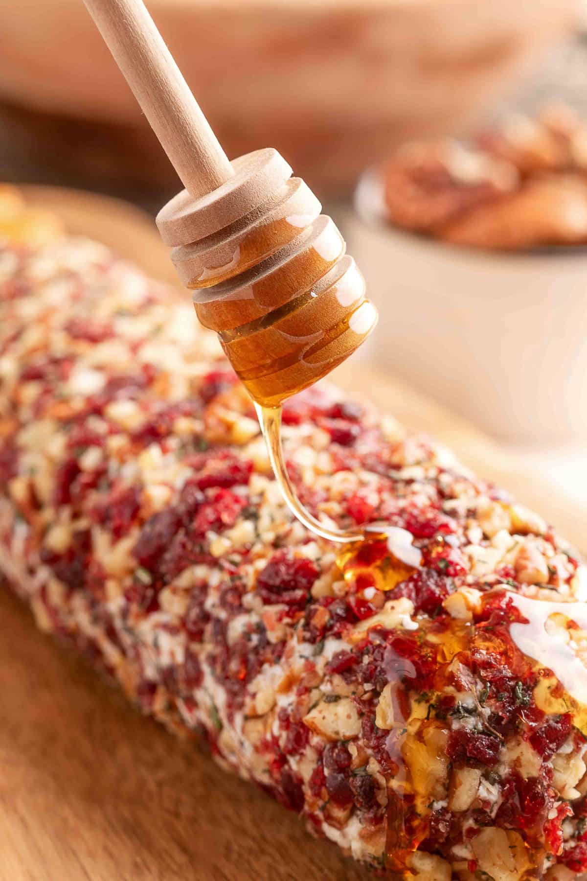 A cranberry pecan-coated goat cheese log resting on plastic wrap atop a wooden cutting board, with scattered bits of the coating and a white bowl in the corner.