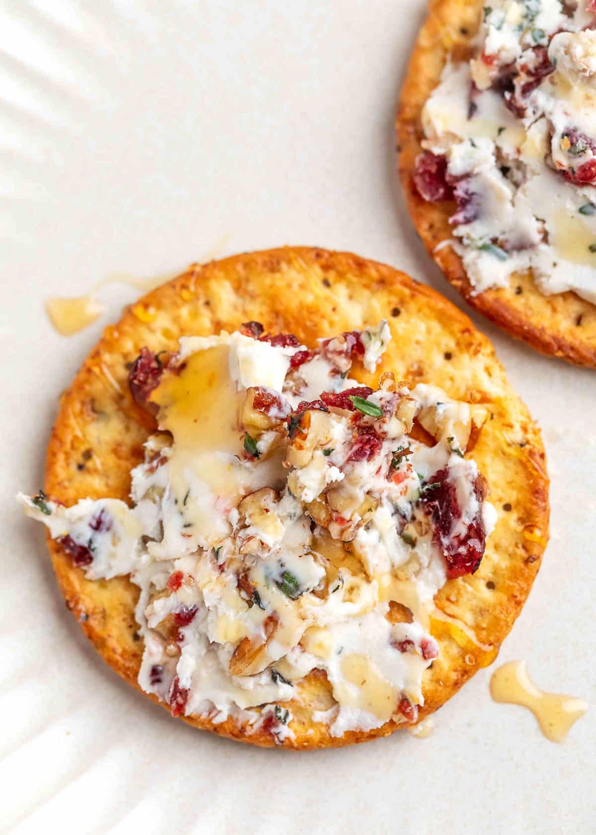 Close-up of a seeded round cracker topped with pecan cranberry goat cheese log and drizzled with honey, served on a white plate.