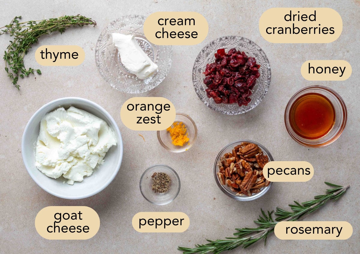 Overhead view of labeled ingredients for a pecan cranberry goat cheese log, including goat cheese, cream cheese, dried cranberries, chopped pecans, honey, orange zest, black pepper, rosemary, and thyme arranged in small bowls and scattered on a light countertop.