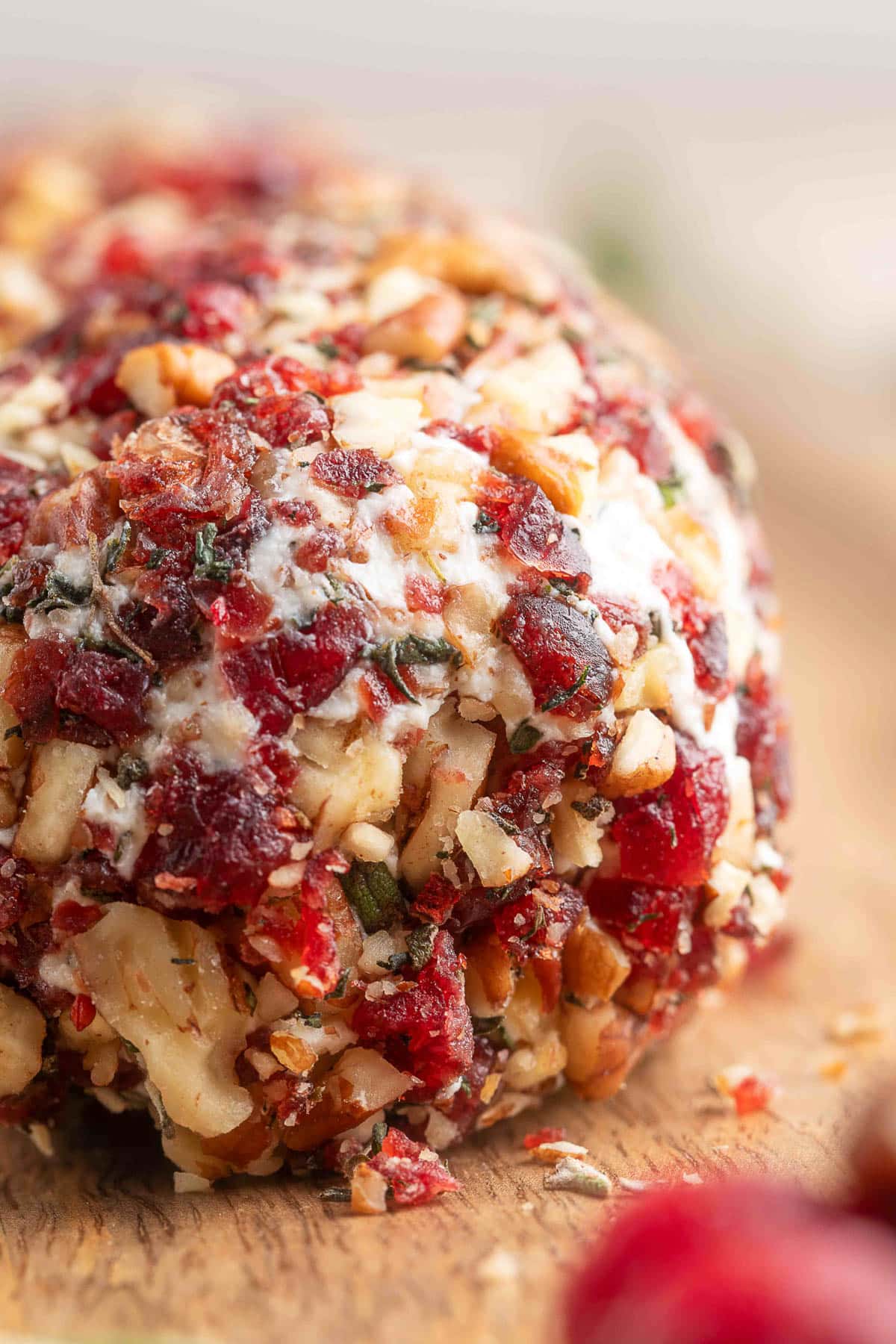Close-up of a creamy goat cheese log sliced open on a wooden board, coated in finely chopped toasted pecans, dried cranberries, fresh rosemary, and thyme, with a drizzle of honey and a sprig of rosemary in the foreground.