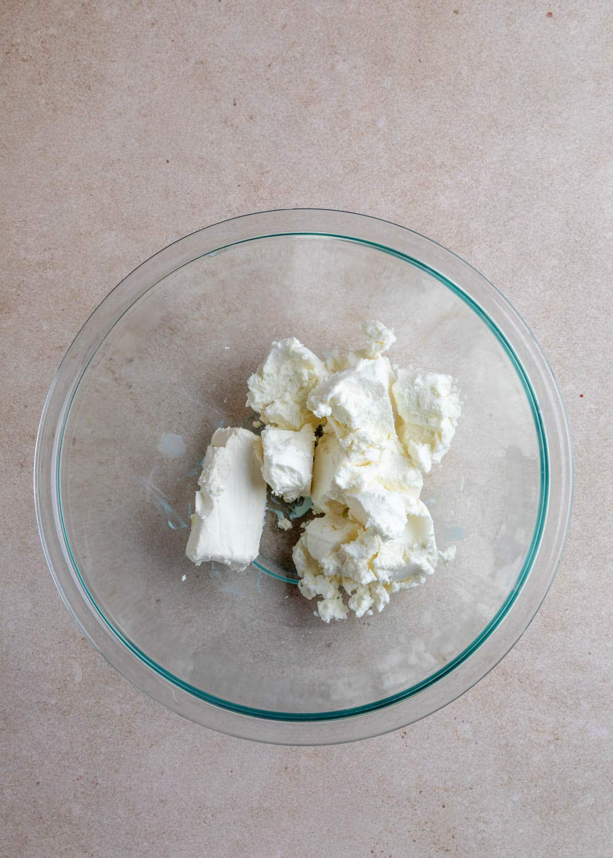 A glass mixing bowl on a neutral stone surface filled with crumbled goat cheese and cream cheese, ready to be blended for a pecan cranberry goat cheese log.