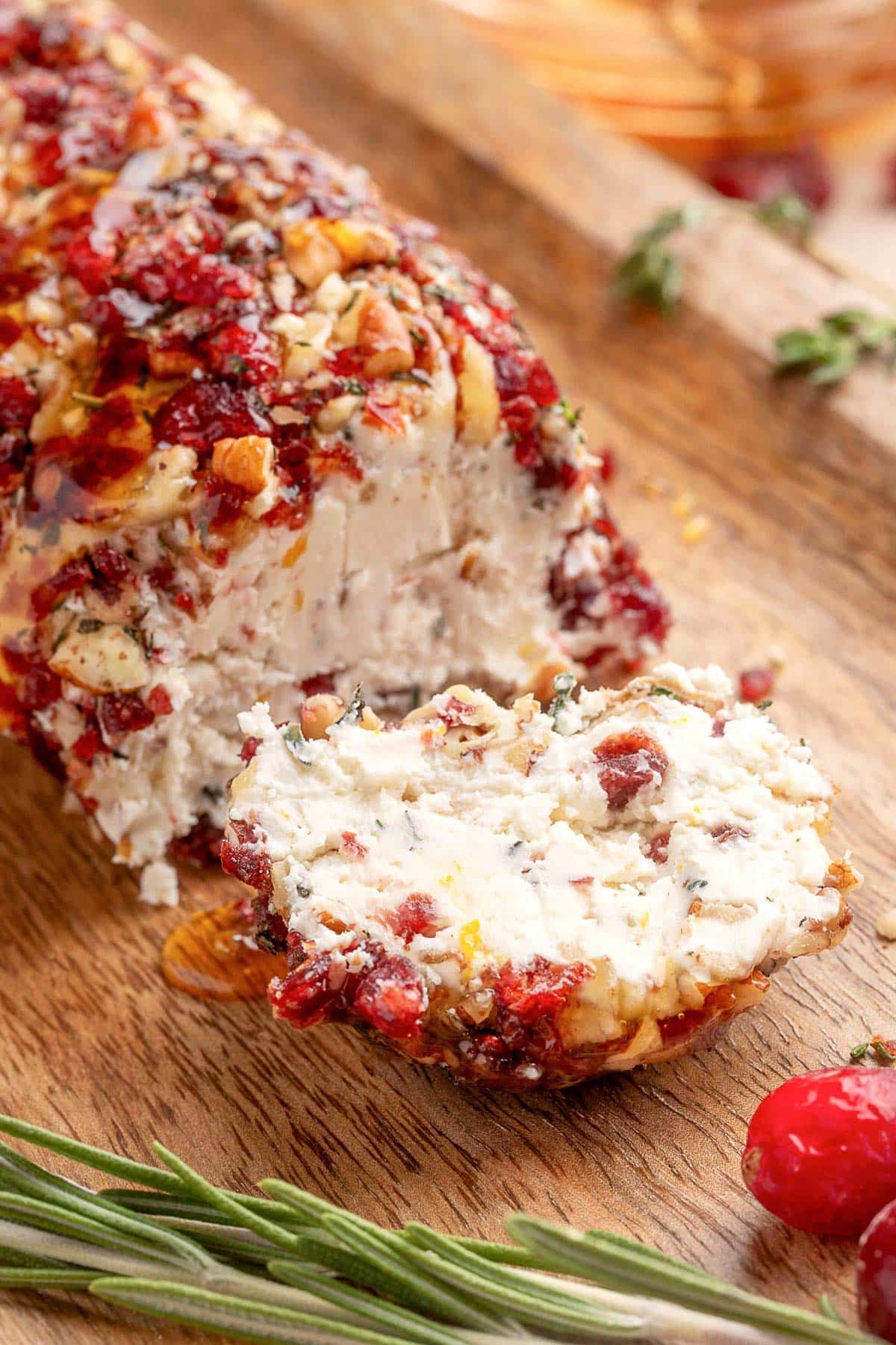 A slice of cranberry pecan goat cheese log resting on a wooden board, with its creamy interior and festive coating of chopped cranberries, toasted pecans, and herbs visible. A drizzle of honey glistens on top, with sprigs of rosemary and a fresh cranberry nearby for garnish.