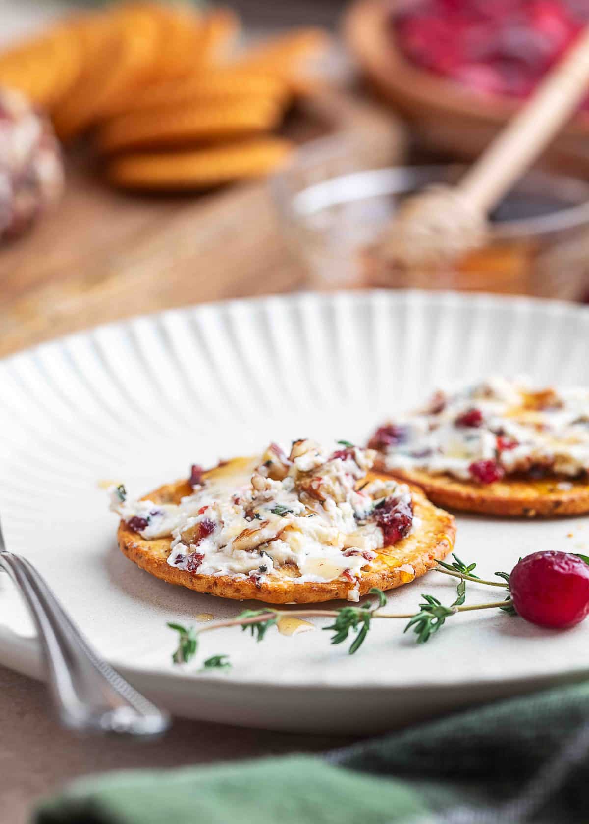 Two golden crackers topped with pecan-cranberry goat cheese log on a white plate, garnished with a sprig of fresh thyme and a whole cranberry, with crackers and a honey dipper blurred in the background.